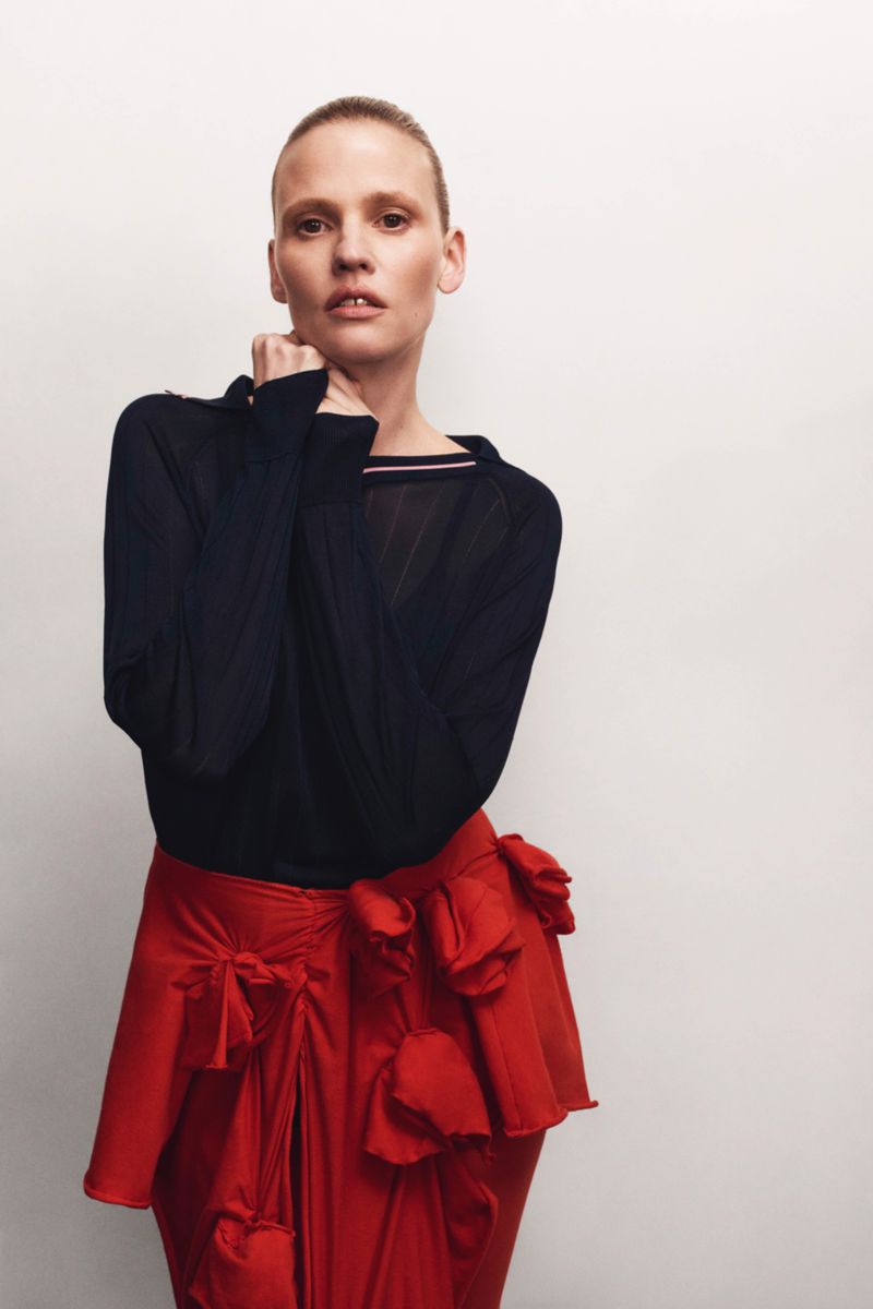 A fair-skinned adult female is posed in front of a plain background wearing a black long-sleeve top and a red skirt with flower-like details.