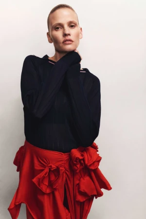 Portrait of a woman with short hair wearing a dark top and a red skirt, standing in front of a white background, looking at the camera.