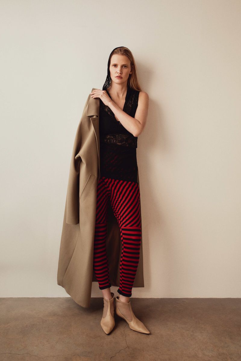 A full shot of a fair-skinned woman wearing a black lace top and red-and-black striped leggings, holding a tan coat over her shoulder.