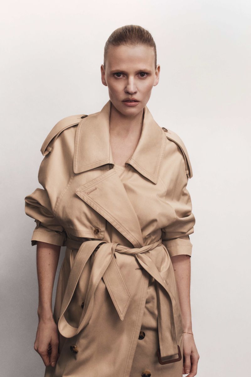 A blond woman poses in a tan trench coat against a neutral backdrop, offering a direct gaze in this fashion-forward studio portrait.