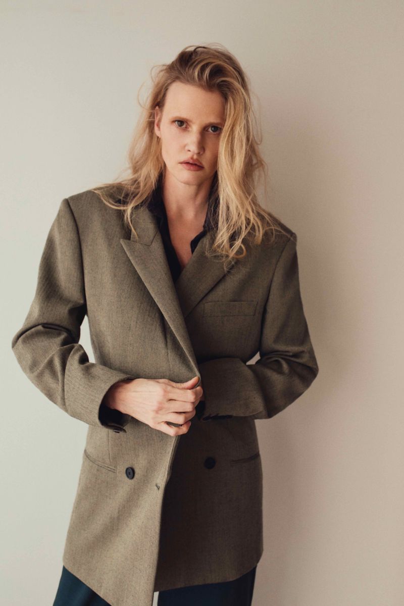 A blonde adult female poses indoors wearing an oversized blazer with long hair and a neutral expression, set against a plain wall.