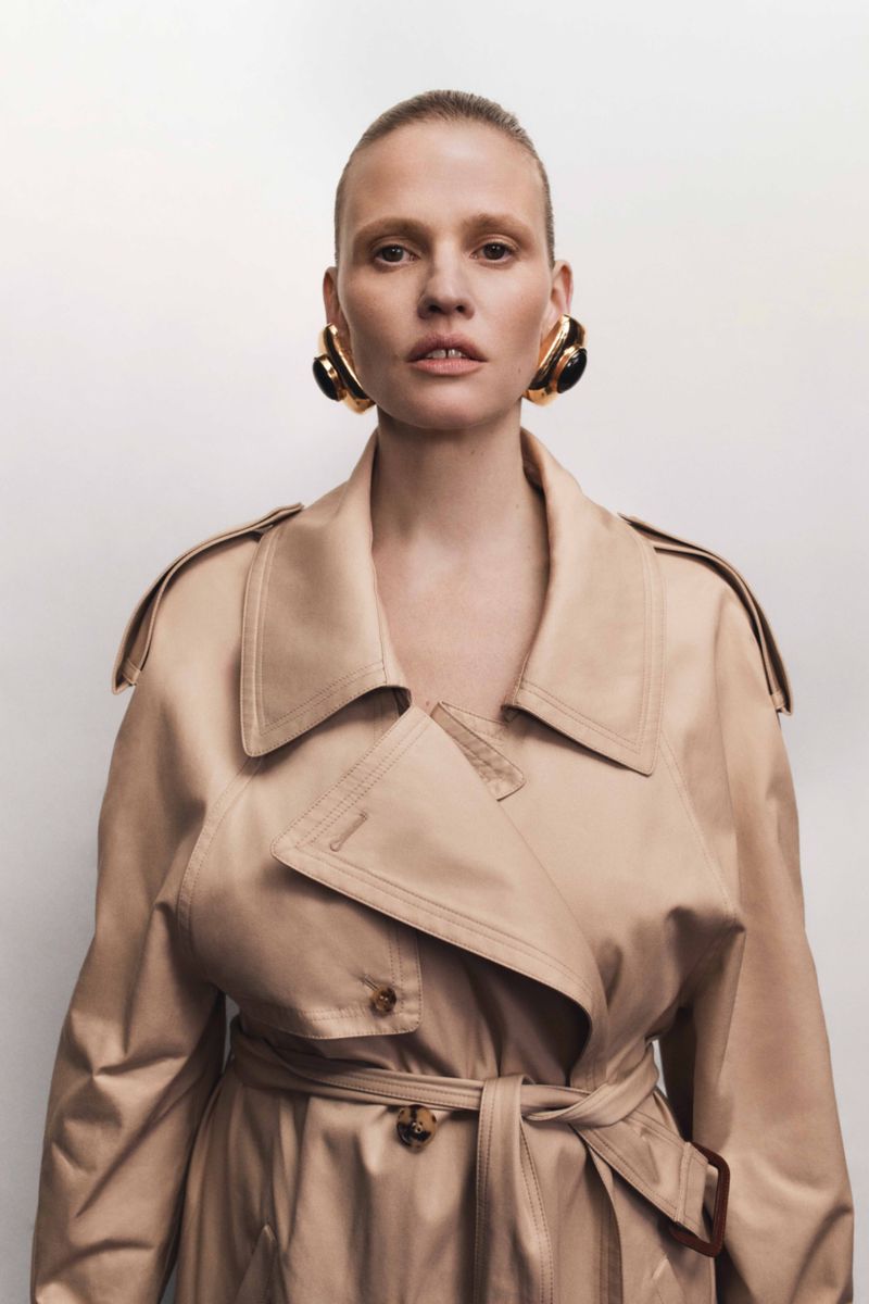 Fashion portrait of a woman with bare skin wearing a beige trench coat and chunky gold earrings against a white backdrop.