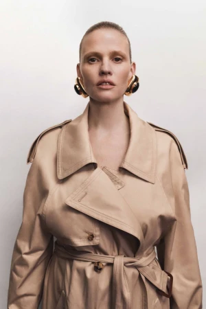 Fashion portrait of a woman with bare skin wearing a beige trench coat and chunky gold earrings against a white backdrop.