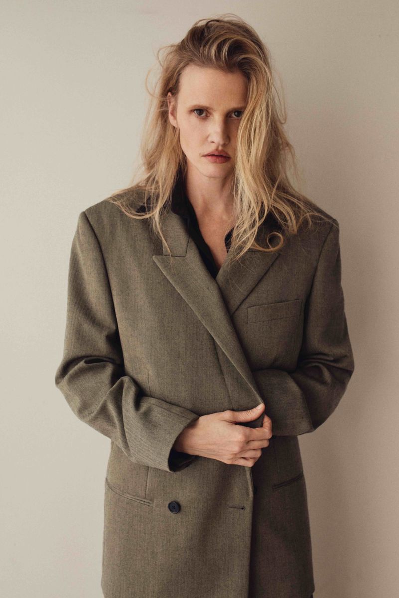 Woman in oversized blazer poses indoors. Portrait shot of blonde adult female in olive-toned herringbone jacket against neutral backdrop.
