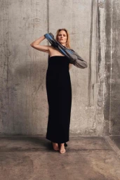 Model Lara Stone wearing a black strapless dress against a textured concrete wall holding a jacket