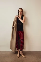 Model Lara Stone in black top and red striped leggings holding camel coat against seamless studio backdrop