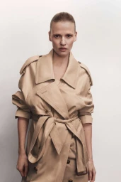 Model Lara Stone wearing a belted beige trench coat against a plain white studio background.