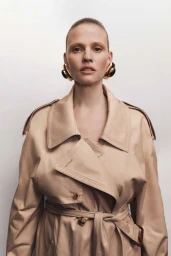 Model Lara Stone wearing a beige trench coat and sculptural earrings against a plain white background