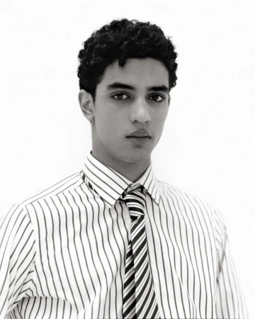 Monochrome portrait of a young adult male with curly hair, wearing a striped shirt and tie, against a bright white backdrop.