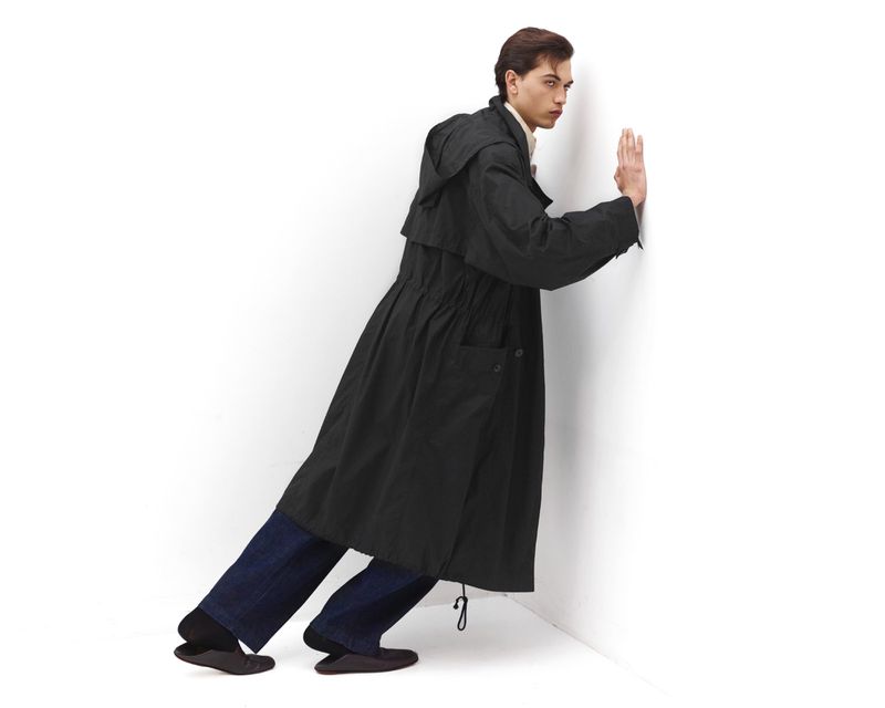 Full-length studio shot of a slender adult man posing in a black hooded trench coat, dark-wash jeans, and dark flats against a white wall.