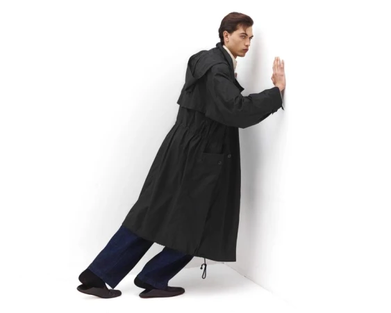 Full-length studio shot of a slender adult man posing in a black hooded trench coat, dark-wash jeans, and dark flats against a white wall.