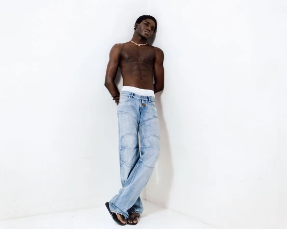 Shirtless black male model wearing light blue denim jeans leaning against a plain white studio corner wall.