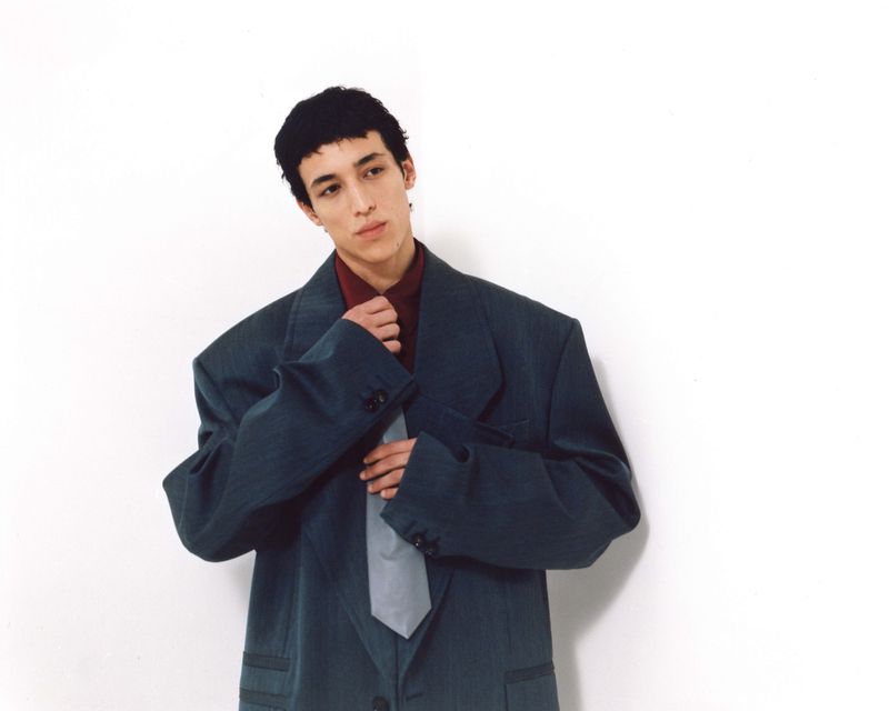 Portrait of a young adult male in an oversized dark teal suit with a grey tie adjusting his collar against a white backdrop.