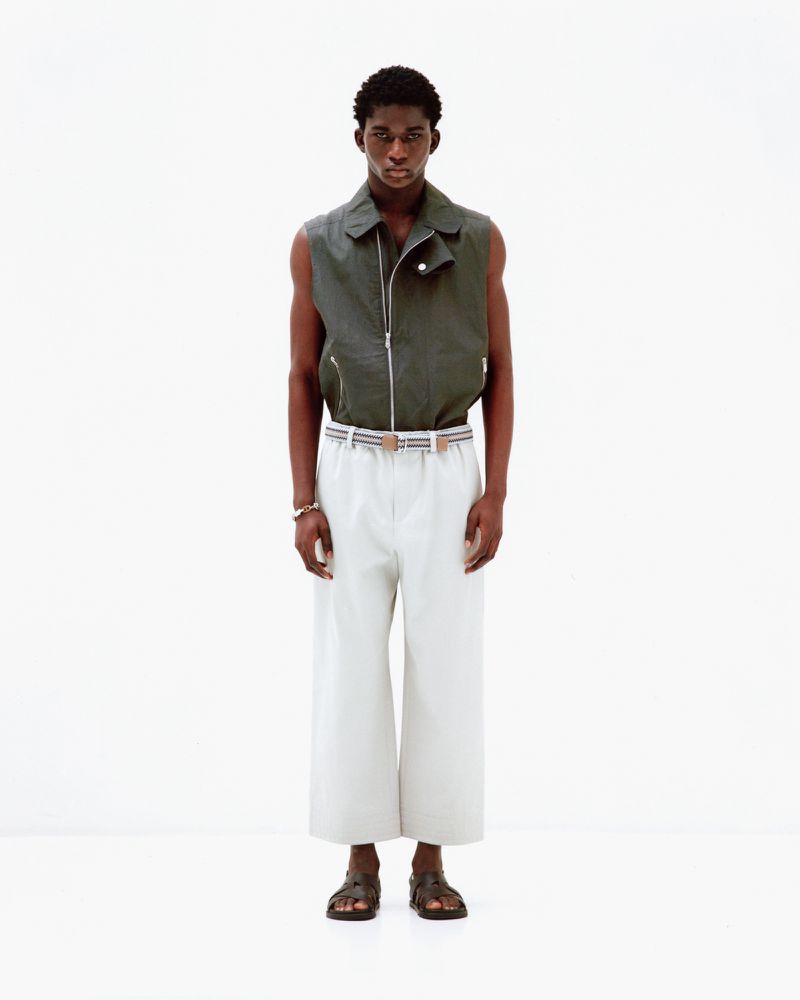 A male model wearing a dark olive sleeveless vest and white trousers standing against a plain white background.
