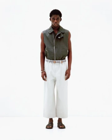 A male model wearing a dark olive sleeveless vest and white trousers standing against a plain white background.