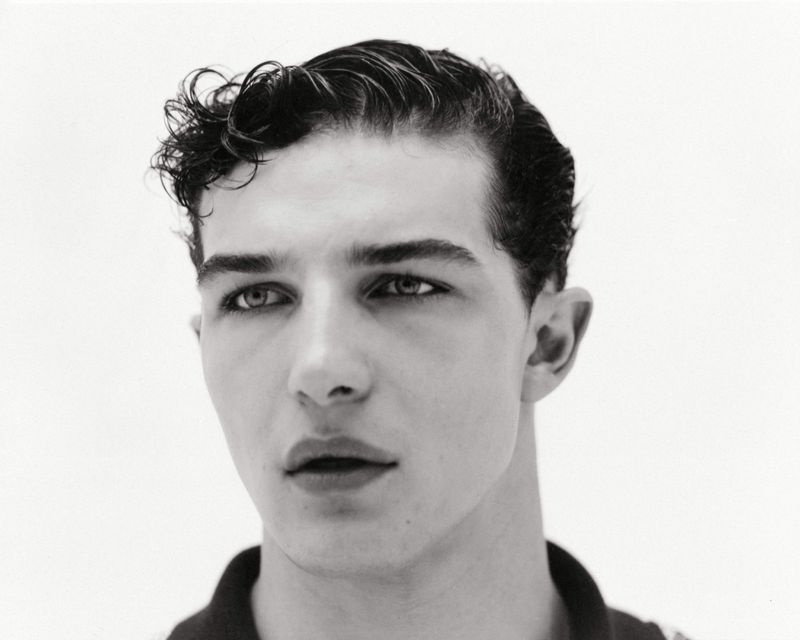 Black and white portrait of a young adult male with dark curly hair, fair skin, and dark eyes, posing against a plain white background.