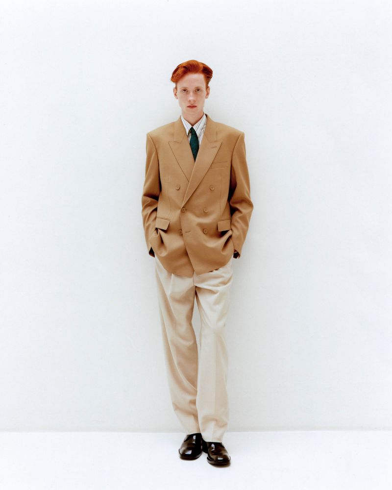 Full-length portrait of a red-haired male model in a tan double-breasted blazer, light trousers, dark tie, and black dress shoes against white.