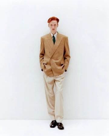Full-length portrait of a red-haired male model in a tan double-breasted blazer, light trousers, dark tie, and black dress shoes against white.