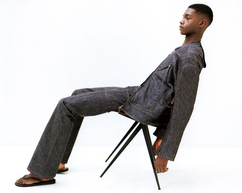 Young adult Black male seated on a stool in a photo studio wearing a raw denim jacket and pants with black flip-flops in studio lighting.