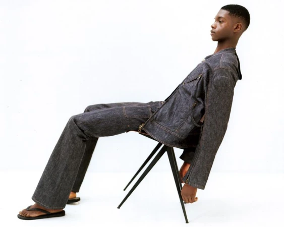 Young adult Black male seated on a stool in a photo studio wearing a raw denim jacket and pants with black flip-flops in studio lighting.