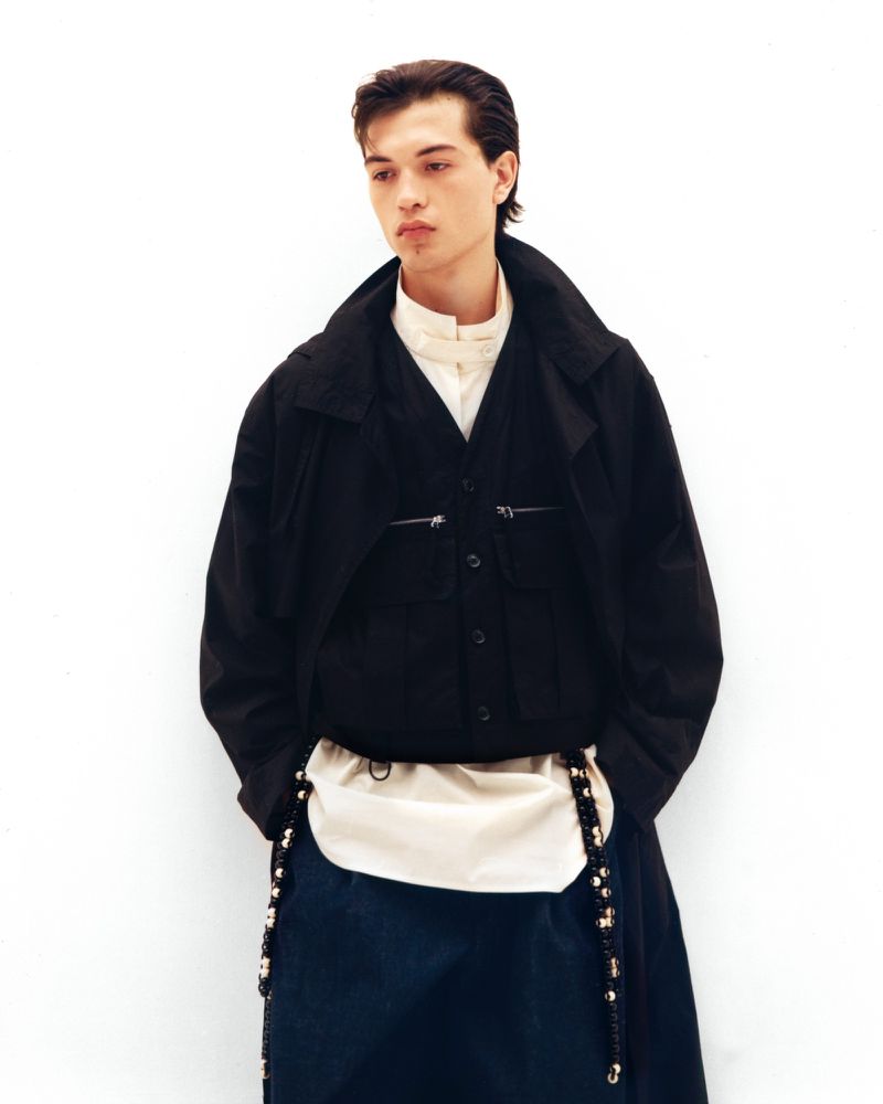 Standing model poses against a white backdrop, wearing a black button-down over a white shirt, and a cream-colored bag with beaded straps.