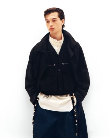 Standing model poses against a white backdrop, wearing a black button-down over a white shirt, and a cream-colored bag with beaded straps.