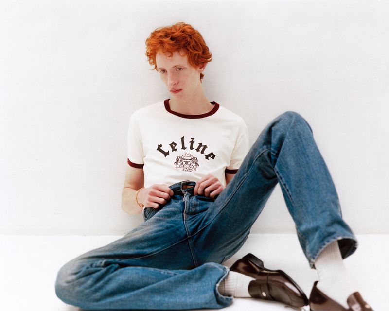 Red-haired male model seated against a white wall wearing a white ringer t-shirt and blue jeans