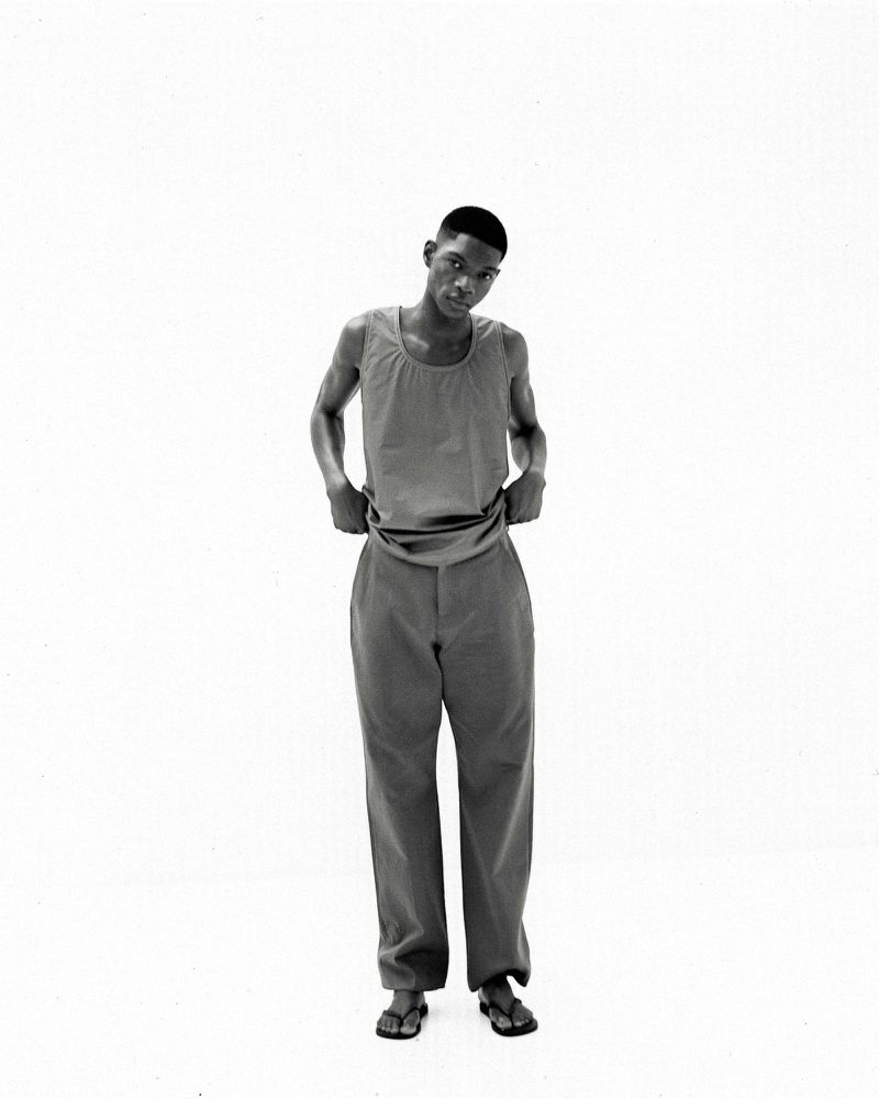 Black and white studio shot of a young, slender Black man wearing a tank top and loose pants, standing barefoot against a plain white backdrop.