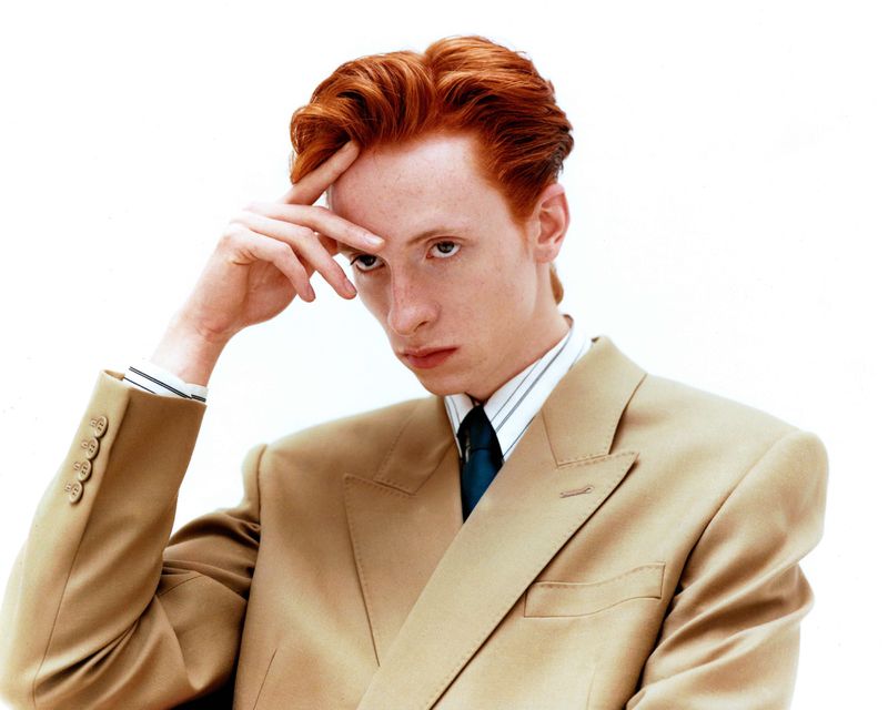 Portrait of a man with red hair in a tan suit looking at the camera with his hand on his forehead against a white background.