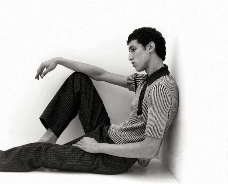 Young man in a textured polo shirt and dark trousers sitting against a white corner wall.