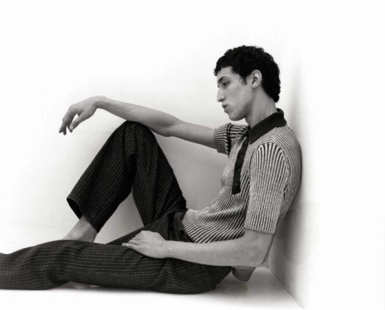 Young man in a textured polo shirt and dark trousers sitting against a white corner wall.