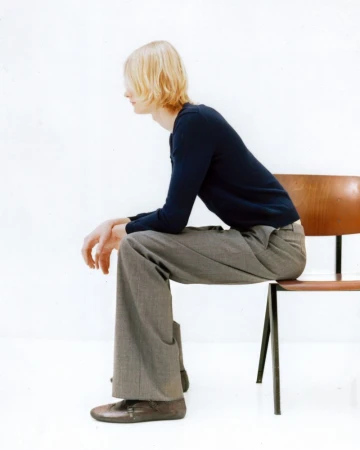 Model with blonde hair seated on a wooden chair wearing a dark sweater and grey trousers against a white background