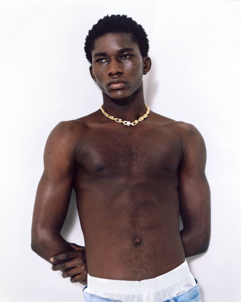 Portrait of a shirtless African American man with a gold chain, standing against a white wall in a studio setting with bright lighting.