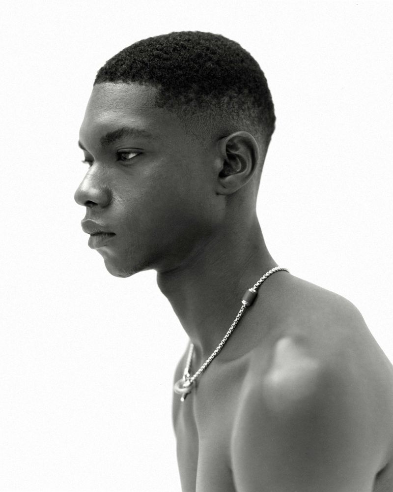 Striking black and white portrait of a young man with short hair, clean-shaven, wearing a silver necklace against a bright white backdrop.