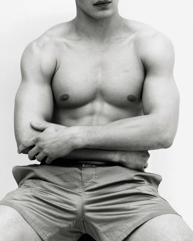 Torso portrait of a shirtless muscular male model with arms crossed wearing shorts against a white background.