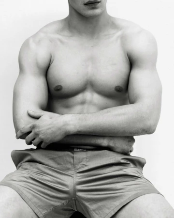 Torso portrait of a shirtless muscular male model with arms crossed wearing shorts against a white background.