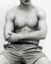 Torso portrait of a shirtless muscular male model with arms crossed wearing shorts against a white background.