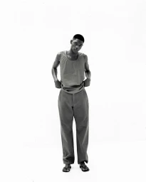 Monochrome full-length portrait of a male model wearing a sleeveless top and trousers against a white seamless backdrop.