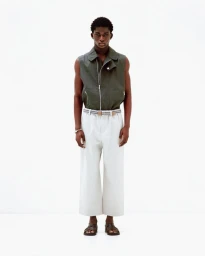 A male model wearing a dark olive sleeveless vest and white trousers standing against a plain white background.
