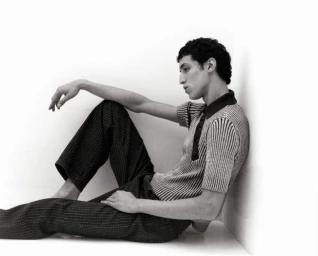 Young man in a textured polo shirt and dark trousers sitting against a white corner wall.
