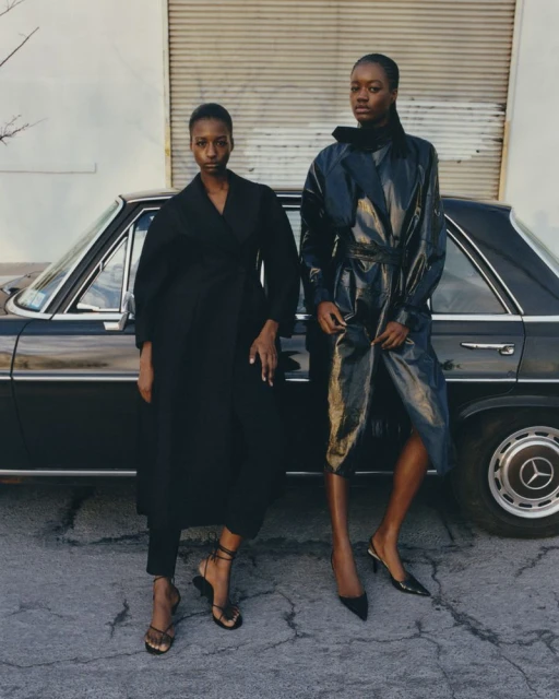 Two Black female fashion models pose in trench coats next to a vintage black car with a building in the background on a cloudy day.