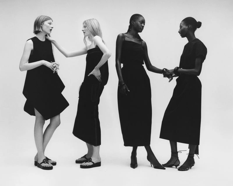 Four fashion models stand together in a studio wearing minimalist black dresses and different styles of shoes in a black and white shot.