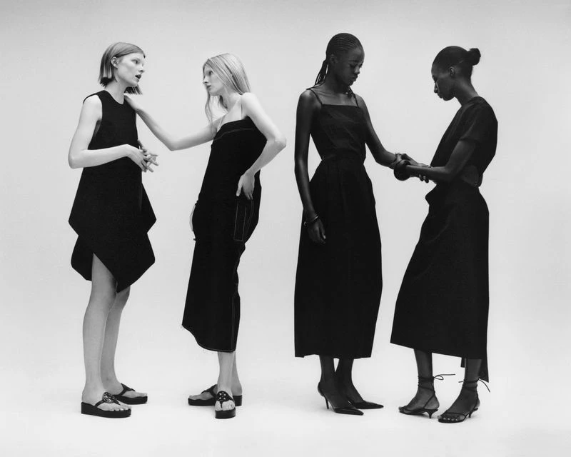 Four fashion models stand in a row against a white backdrop wearing dark, elegant dresses and shoes in a minimalist, studio setting.