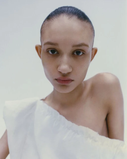 Close-up studio portrait of a young adult female with shaved head, wearing a white fabric drape, against a bright white background.