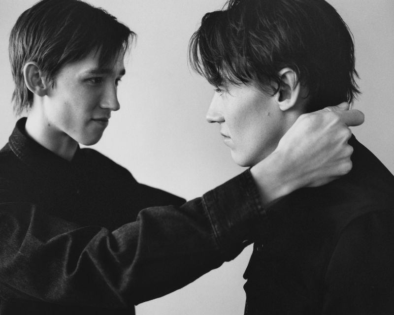 Black and white portrait of two young adult men in dark jackets facing each other in front of a neutral background.