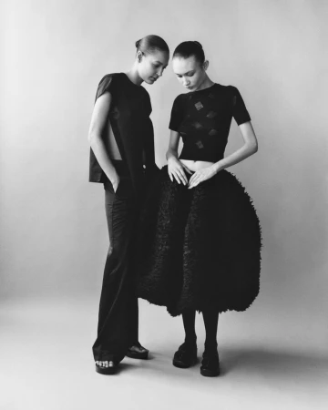 Two women in dark, high-fashion attire pose indoors against a pale backdrop. One wears a boxy top and wide-leg pants, the other a textured dress.