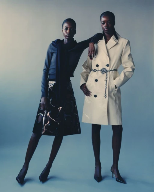 Two Black female fashion models pose in a studio wearing a trench coat and skirt in a medium studio shot with a light-blue backdrop.