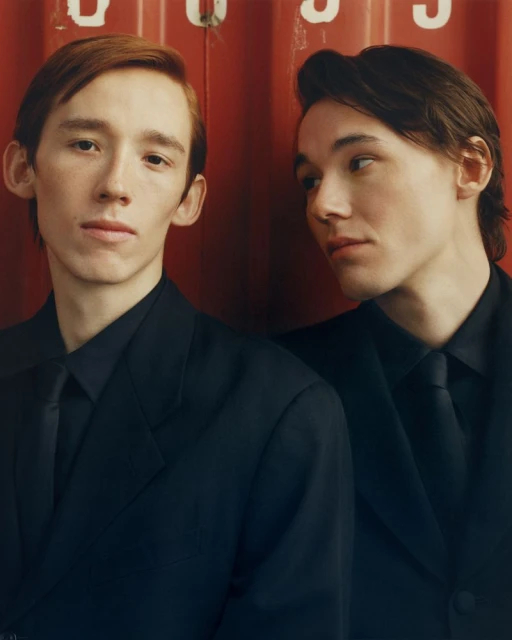 Two young adult men in dark suits and ties pose indoors against a red industrial background, creating a portrait conveying formal style.