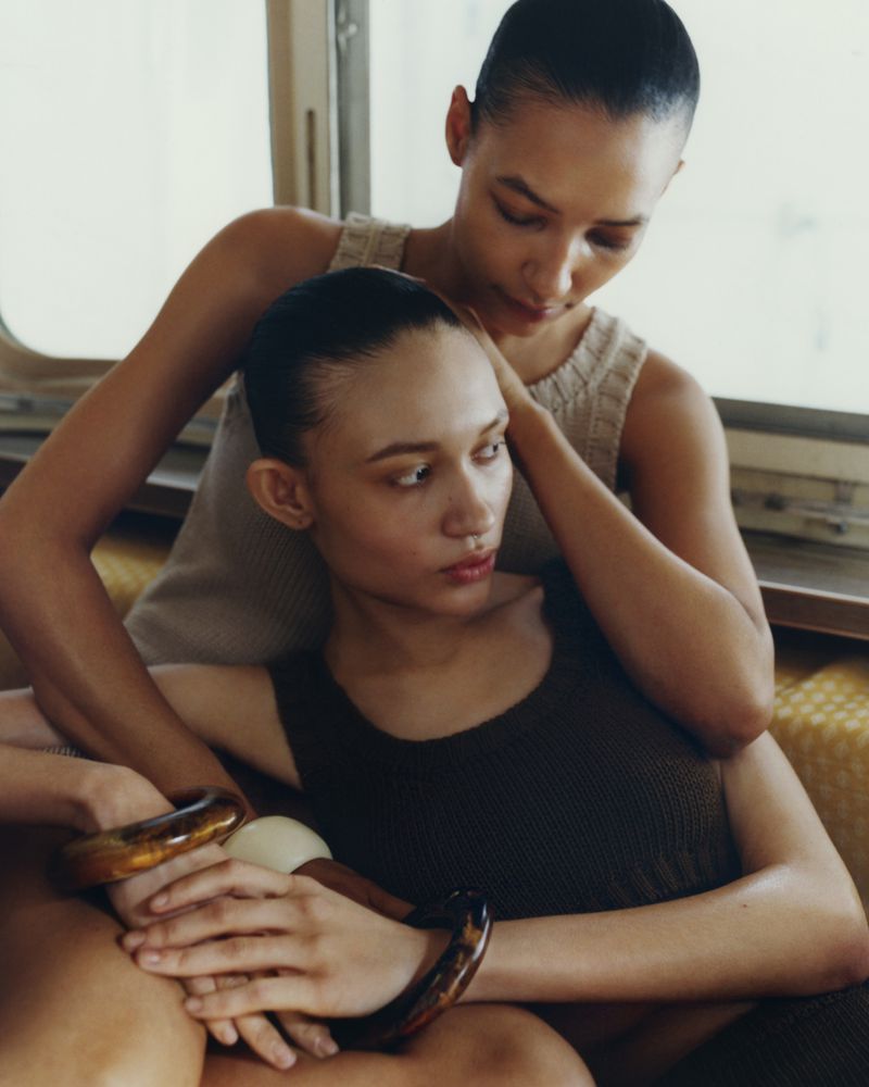 Two women are posing indoors next to a window in a close-up shot. One woman is seated while the other woman is standing behind her.