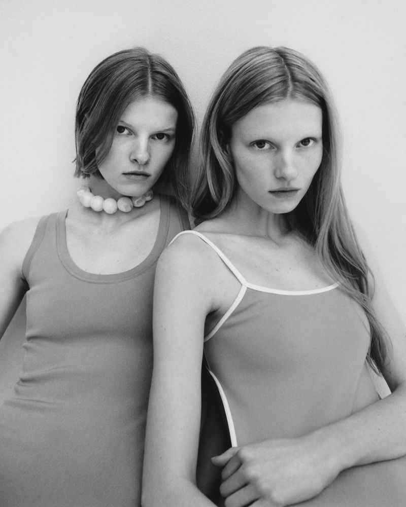 Black and white portrait of two young adult women posing indoors. One wears a sphere necklace, and both are wearing tank tops.